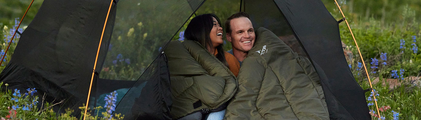 A man and a woman laugh together while snuggled in their Mammoth sleeping bag in a field of wildflowers.