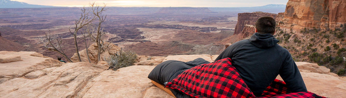 A man sits in his sleeping bag looking out at a scenic view.