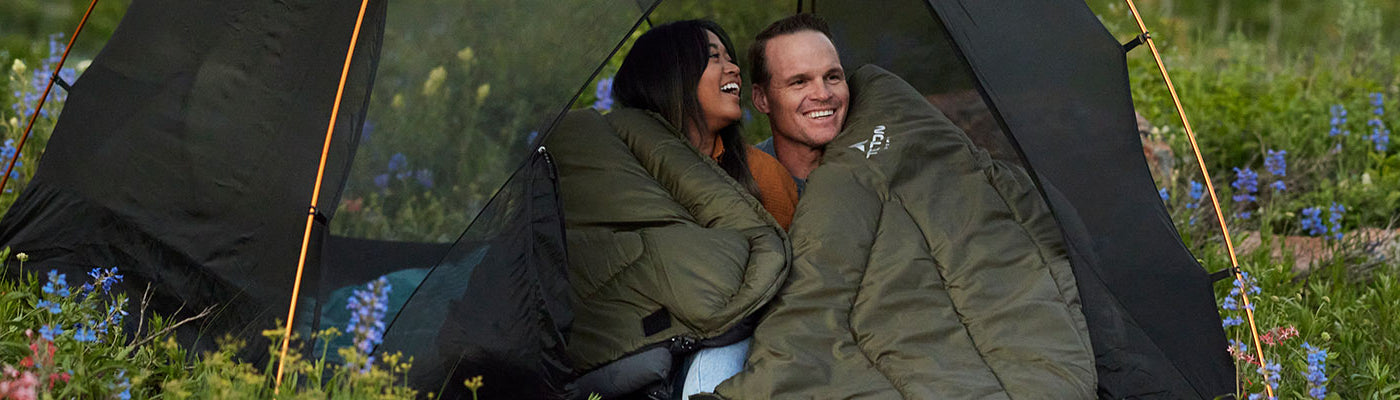 A couple snuggles up in their TETON Sports Mammoth sleeping bag while sitting in a Mountain Ultra tent.