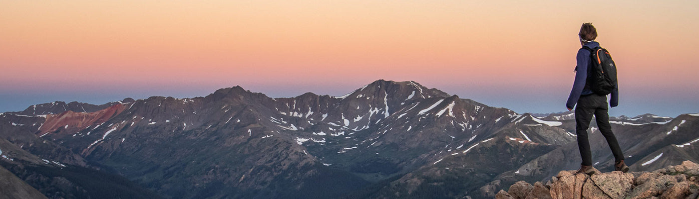 A sole hiker stands atop a rocky acrop surrounded by mountains while wearing a TETON Sports backpack.