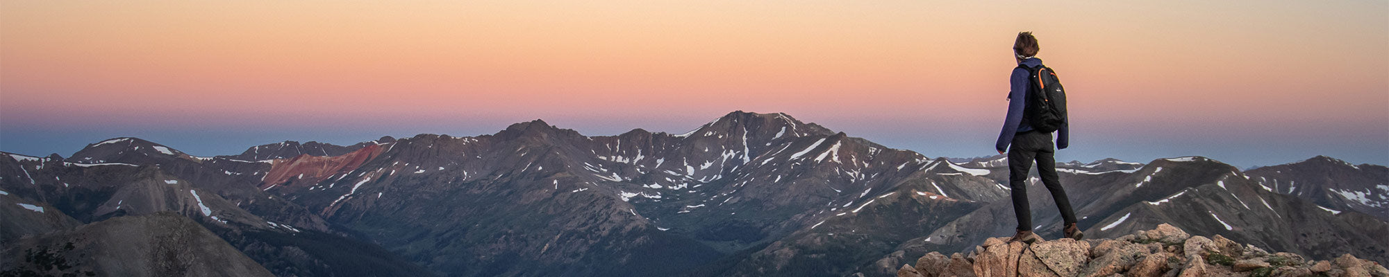 A sole hiker stands atop a rocky acrop surrounded by mountains while wearing a TETON Sports backpack.