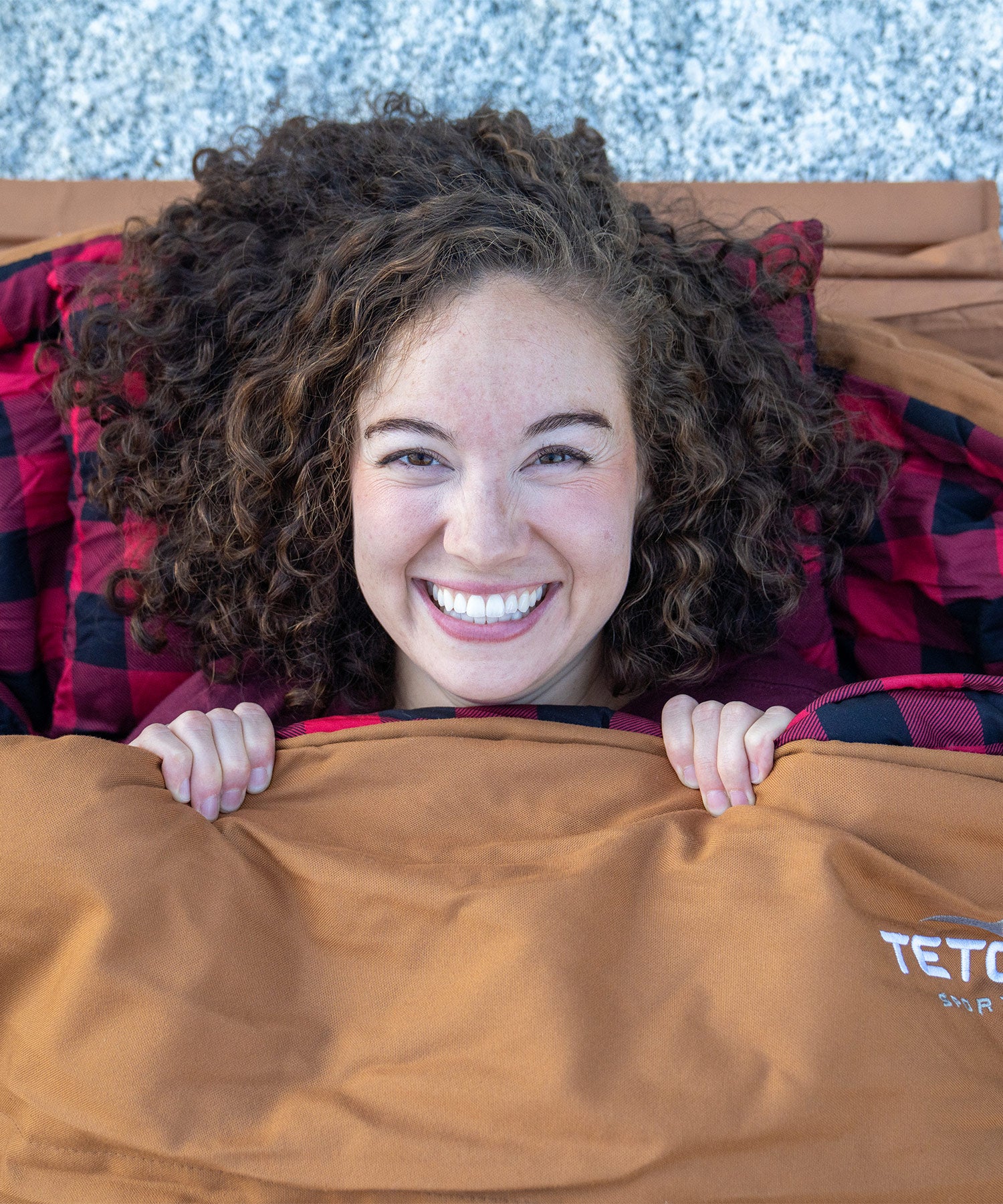 Image shows a woman smiling while snuggled in a brown TETON Sports Bridger Canvas Sleeping Bag.