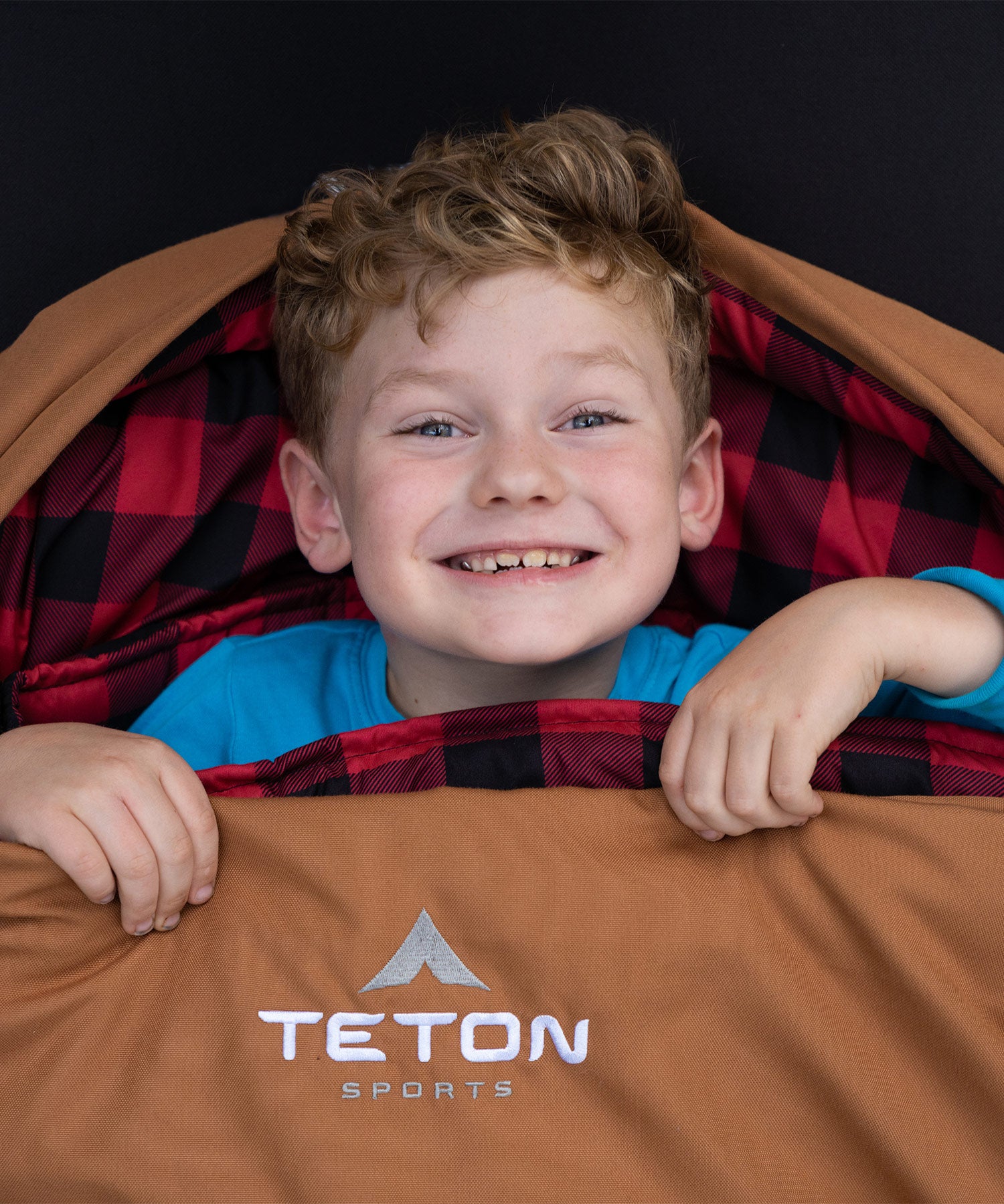 Image shows a young boy smiling while snuggled in a brown TETON Sports Bridger Canvas Sleeping Bag.