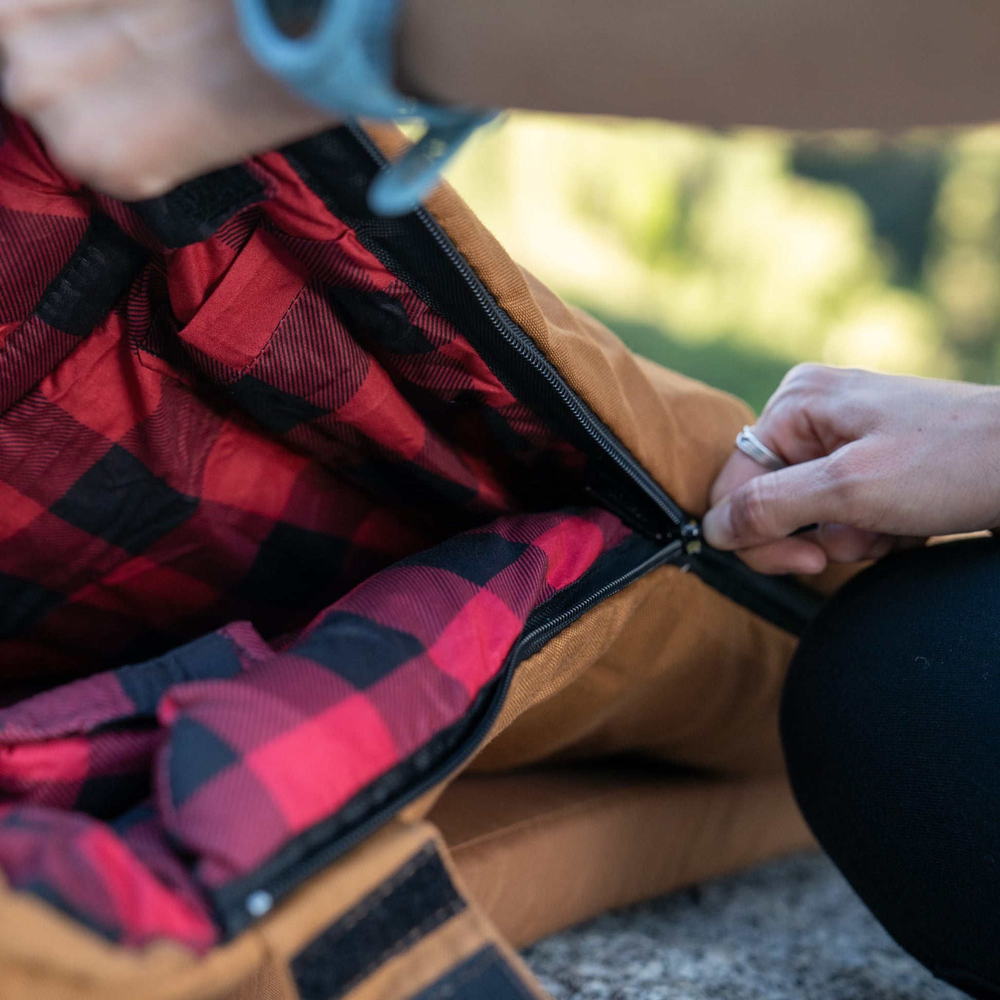 Person opening a red and black checkered sleeping bag with a blurred natural background