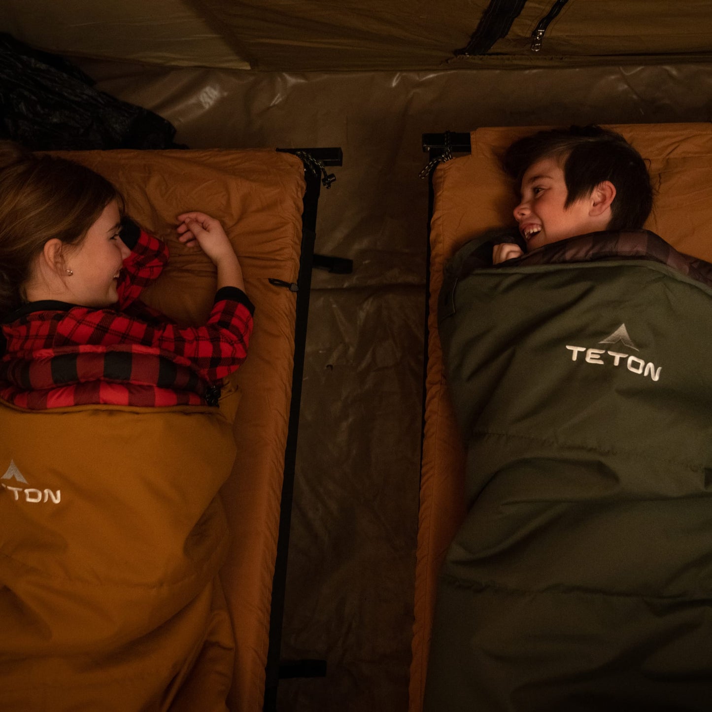 Two people lying in sleeping bags inside a tent, with 'Teton' branding visible on their clothing.