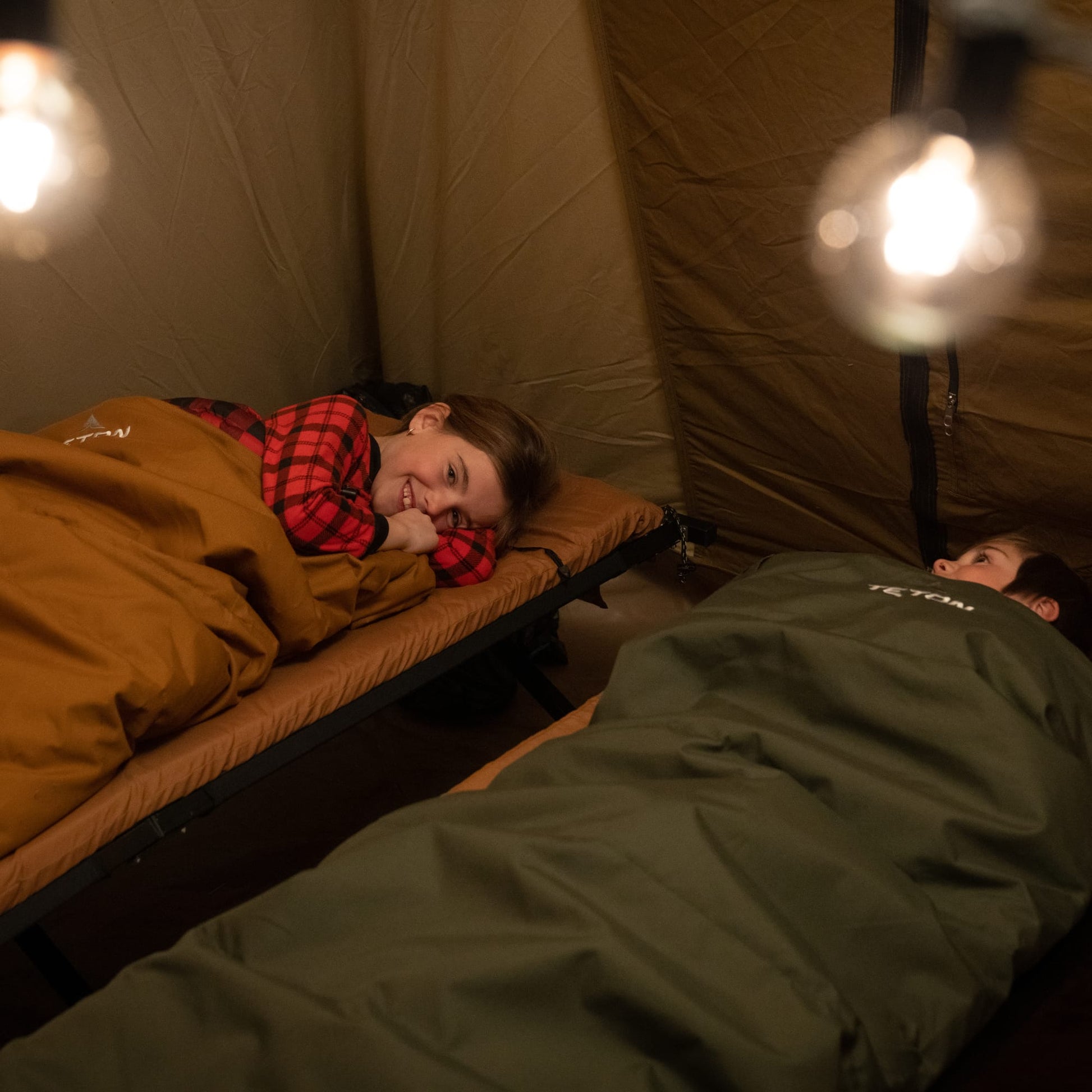 Two kids lying in a bridger sleeping bags inside a tent with warm lighting.