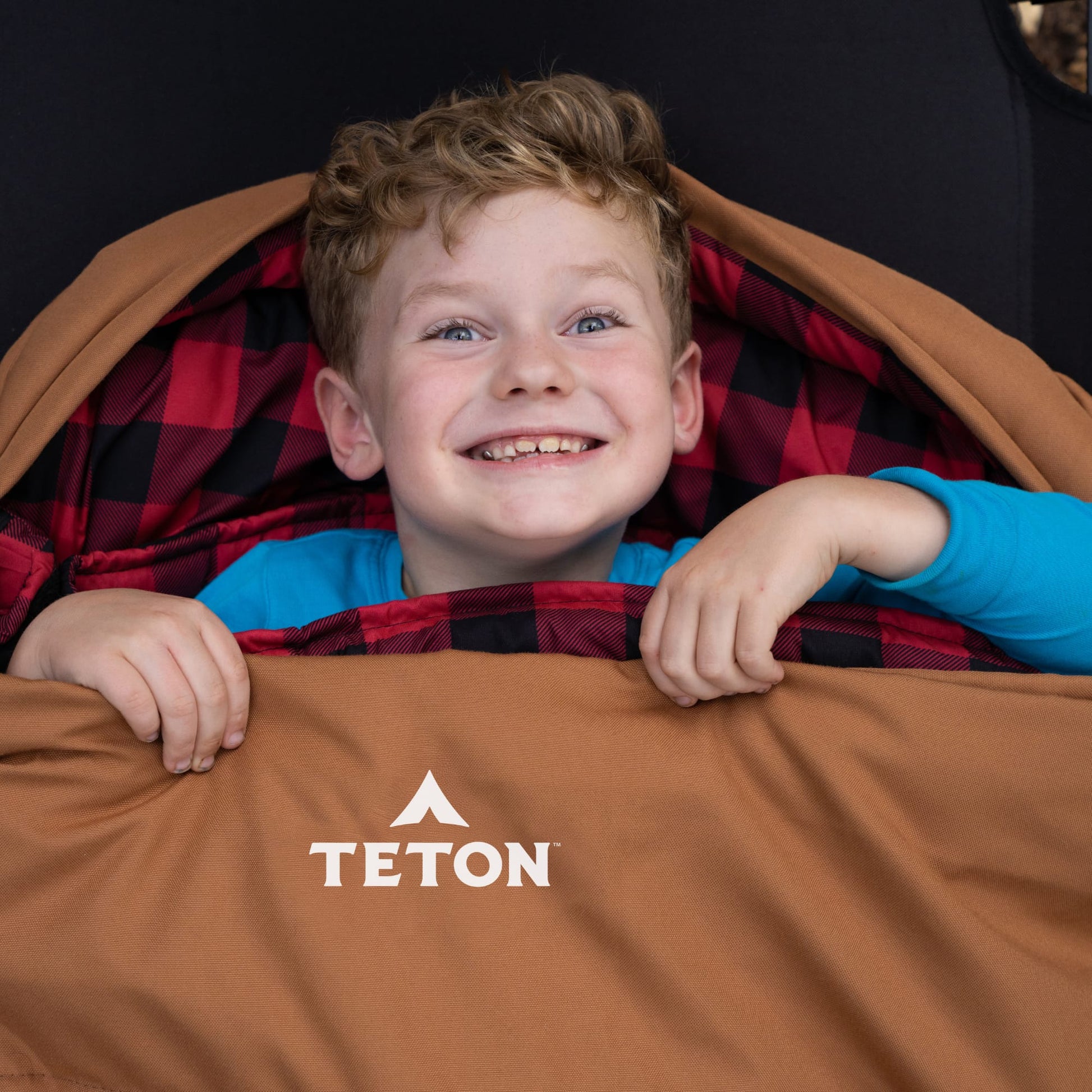 Child peeking out from behind a brown sleeping bag with Teton brand logo
