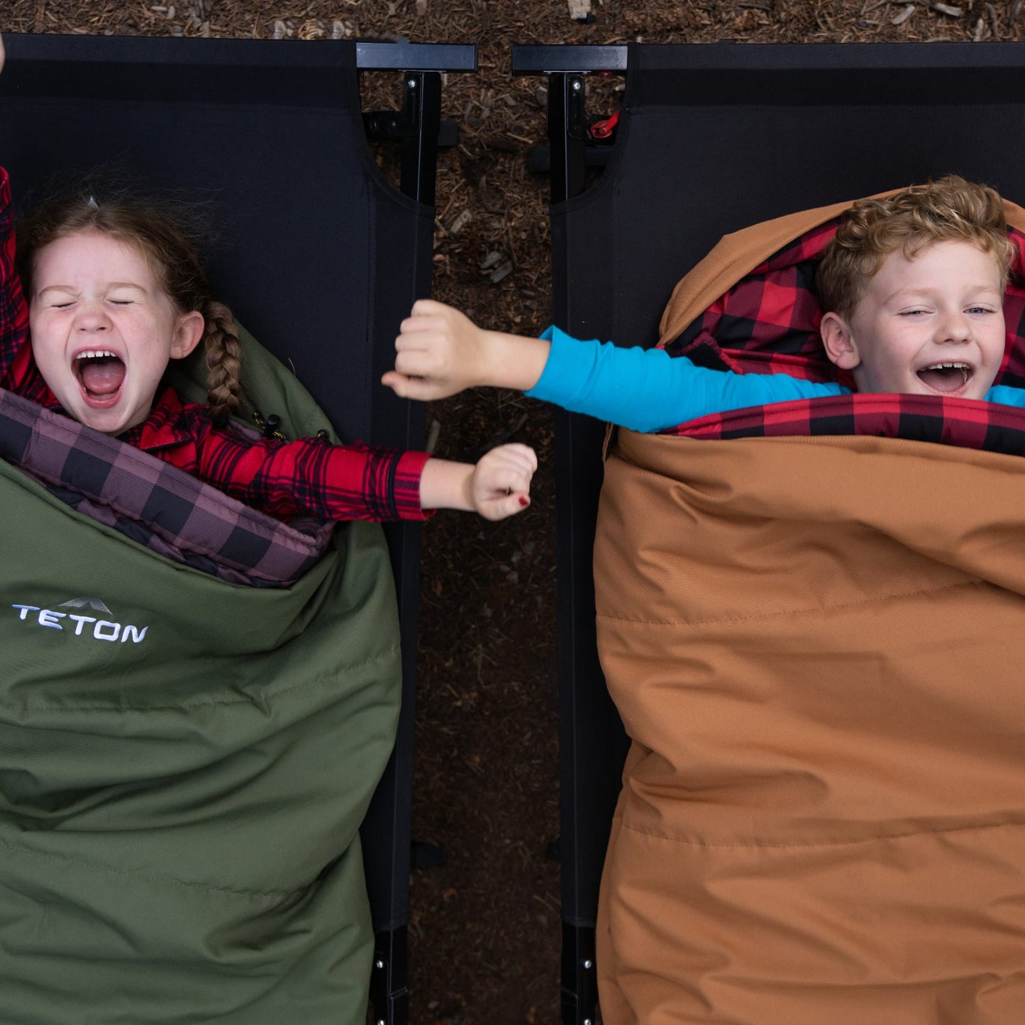 Two children in bridger sleeping bags, one green and one brown, with TETON brand visible, against a natural background.