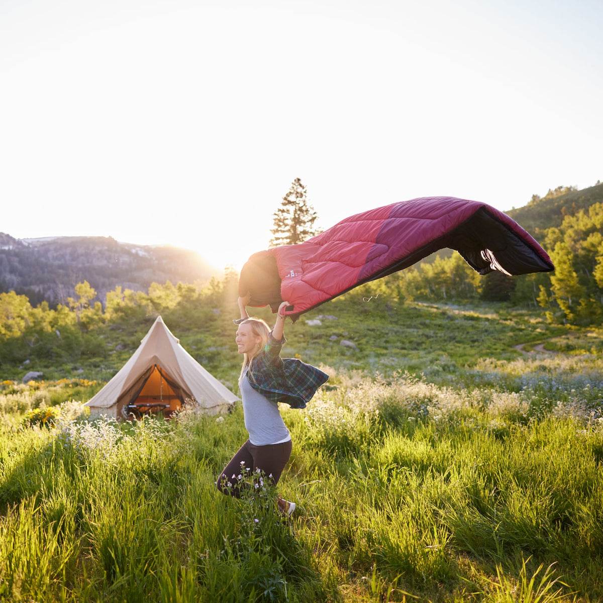 Person holding a Celsius Ruby sleeping bag in a scenic outdoor setting with a tent and mountains.