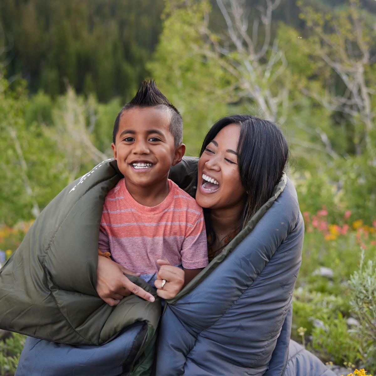 Mother and son people laughing together in a Celsius sleeping bag outdoors with greenery in the background