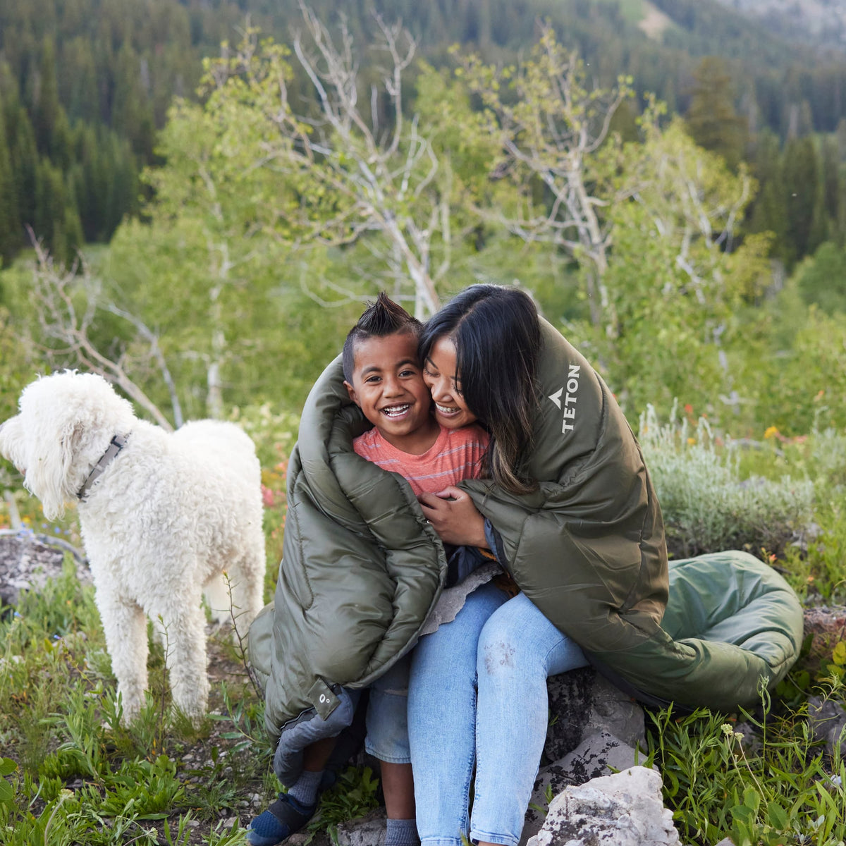 Mother and son sitting on a rock wrapped in a Celsius Sleeping Bag with a white dog in a forested area