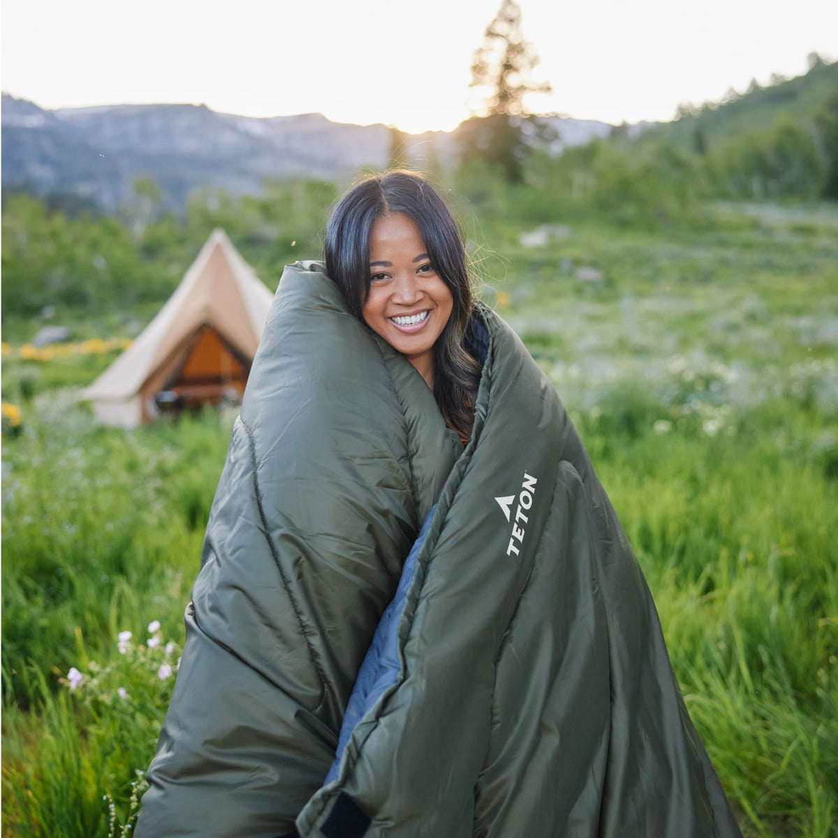 Person wrapped in a celsius sleeping bag in a grassy field with the sierra tent and mountains in the background.