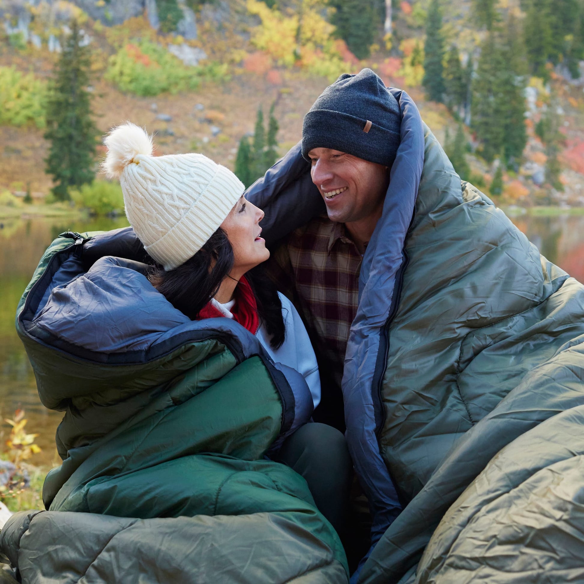 Two people sitting together in a celsius sleeping bags with a scenic background of trees and mountains.
