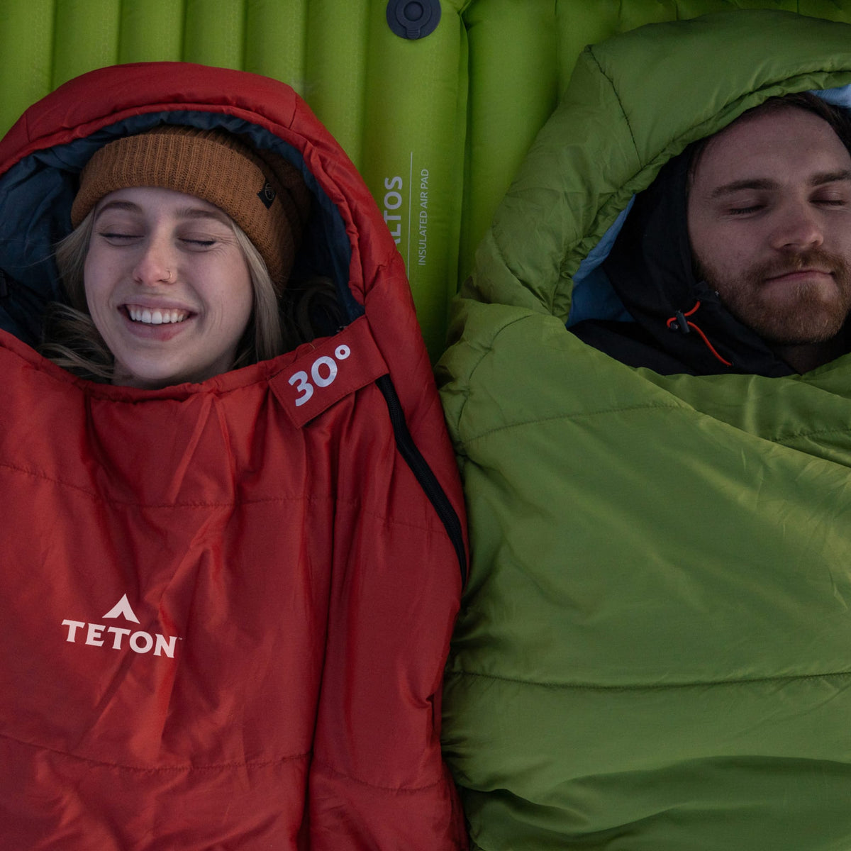 Two people in red and green leef mummy sleeping bags with Teton brand logos, lying on a green sleeping pad.
