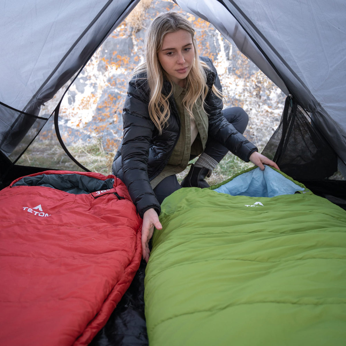 Woman setting up a red and green leef mummy sleeping bag inside a tent with a forest background.