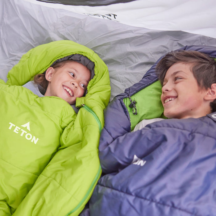 Two children in leef mummy sleeping bags with TETON brand logos, lying inside a tent.