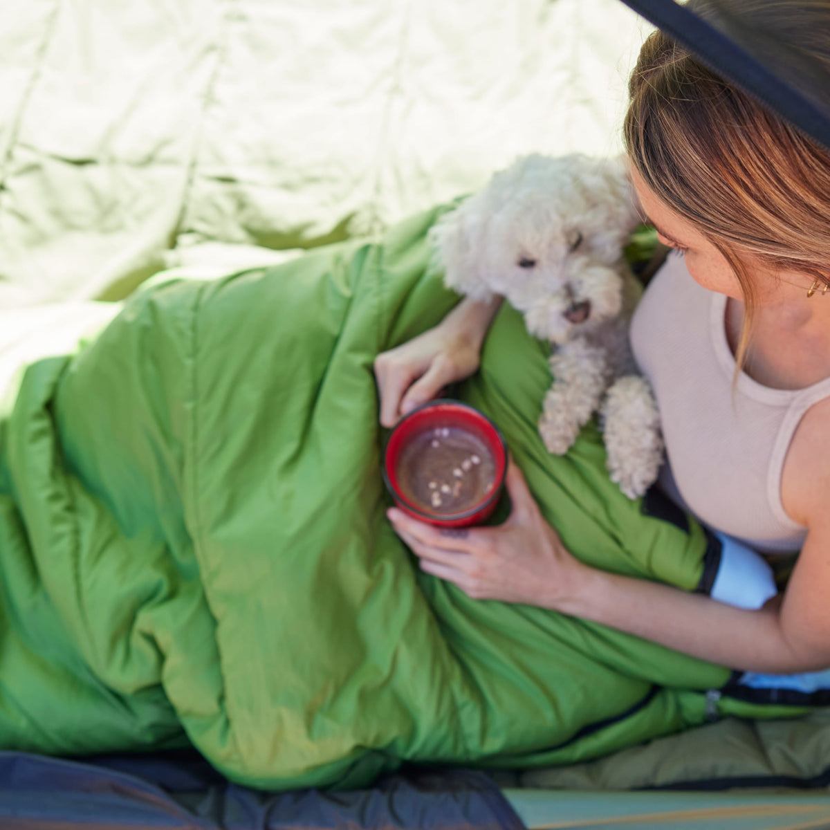 Person lying in a green sleeping bag with a small white dog and a red cup, camping.