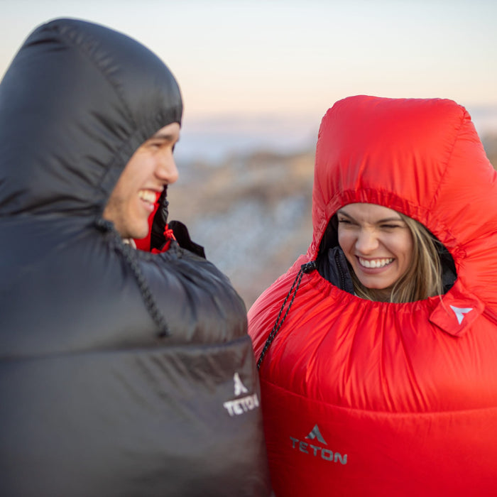 Two people wearing black and red altos mummy sleeping bags with Teton brand logos against a scenic background.