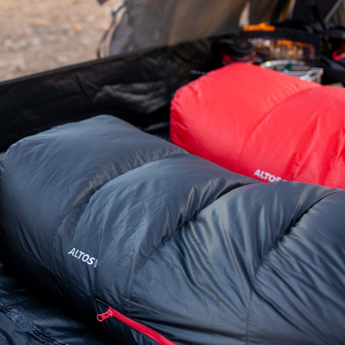 Two altos mummy sleeping bags, one black and one red in a camping setting.