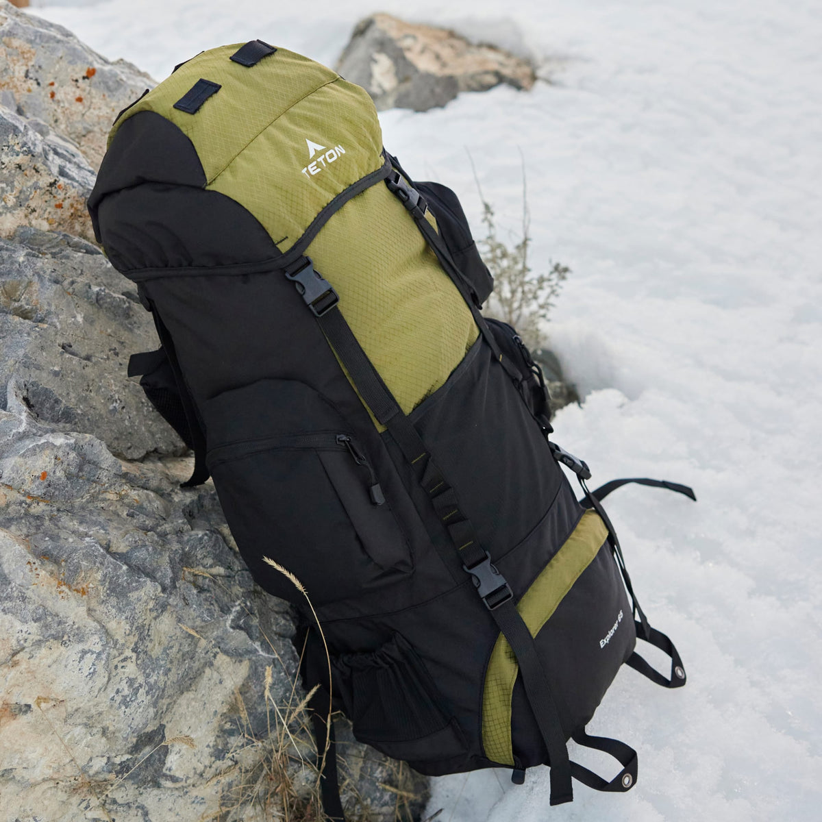 Black and olive green 65L backpack leaning against a rock with snow in the background