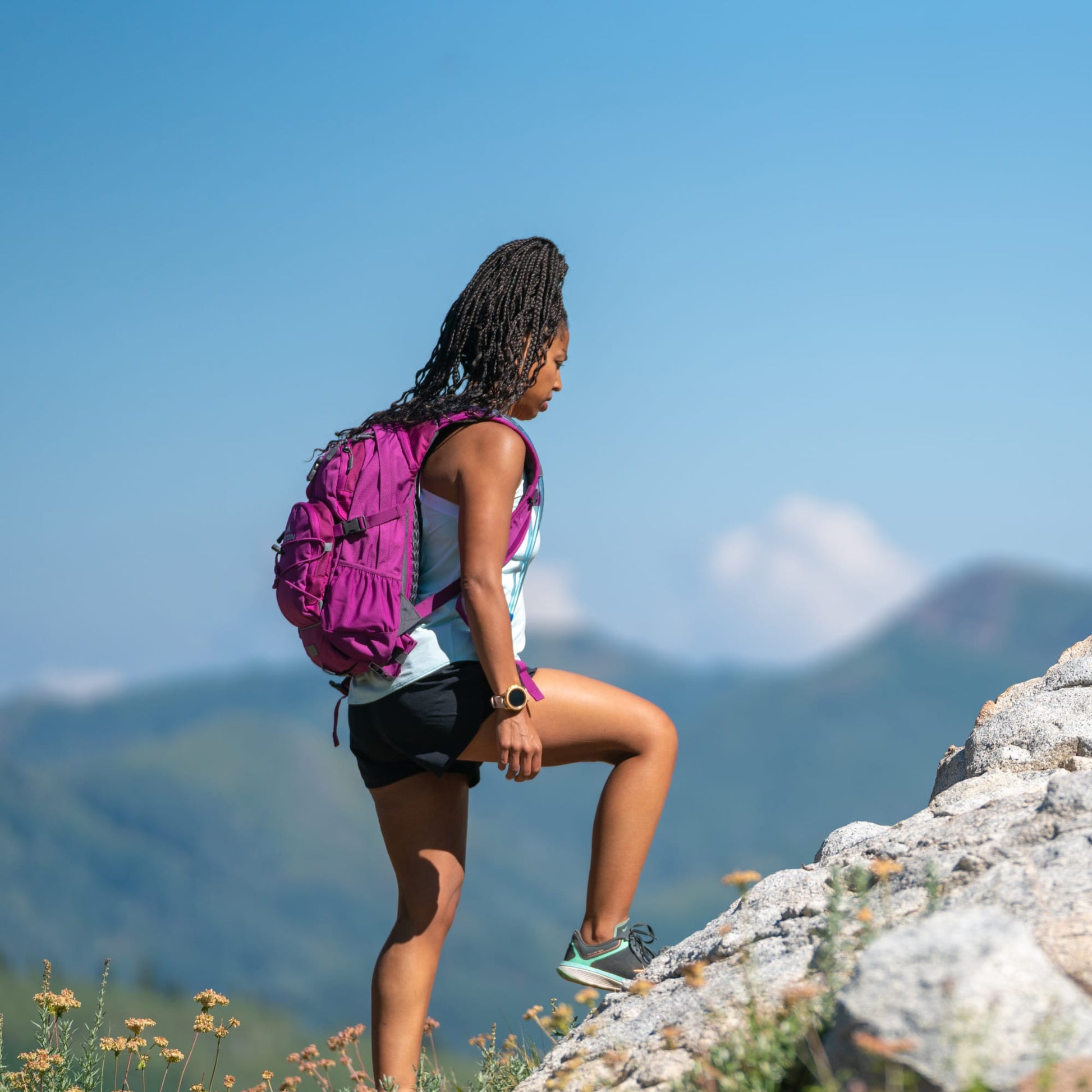Person with a purple hydration backpack standing on a rocky mountain top with a clear blue sky.