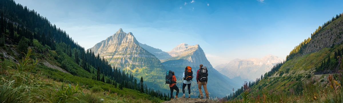 Three hikers with backpacking packs standing on a ridge overlooking a mountain valley at sunrise.