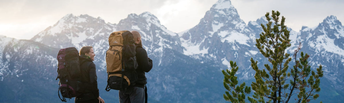 Couple wearing backpacking packs looking out at the snow-capped Teton mountain range.