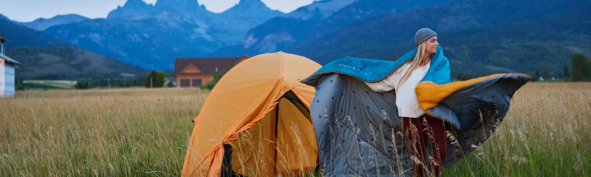 Woman holding open a teal and goldenrod camp blanket next to an yellow tent, Teton mountains in the background.
