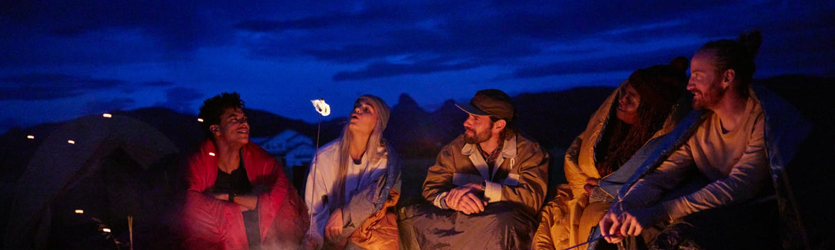 Group of friends wrapped in colorful camp blankets around a campfire at dusk with tents behind them.