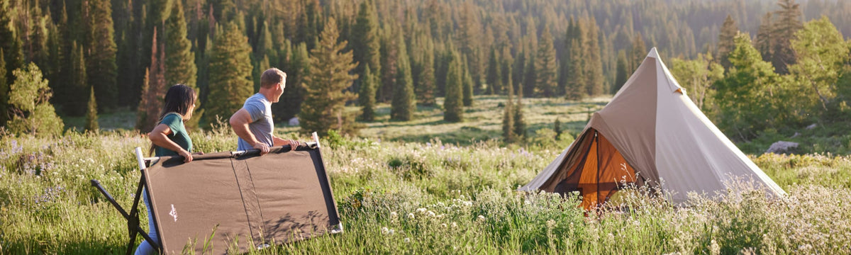 Couple carrying a folded camping cot across a wildflower meadow toward a canvas bell tent.