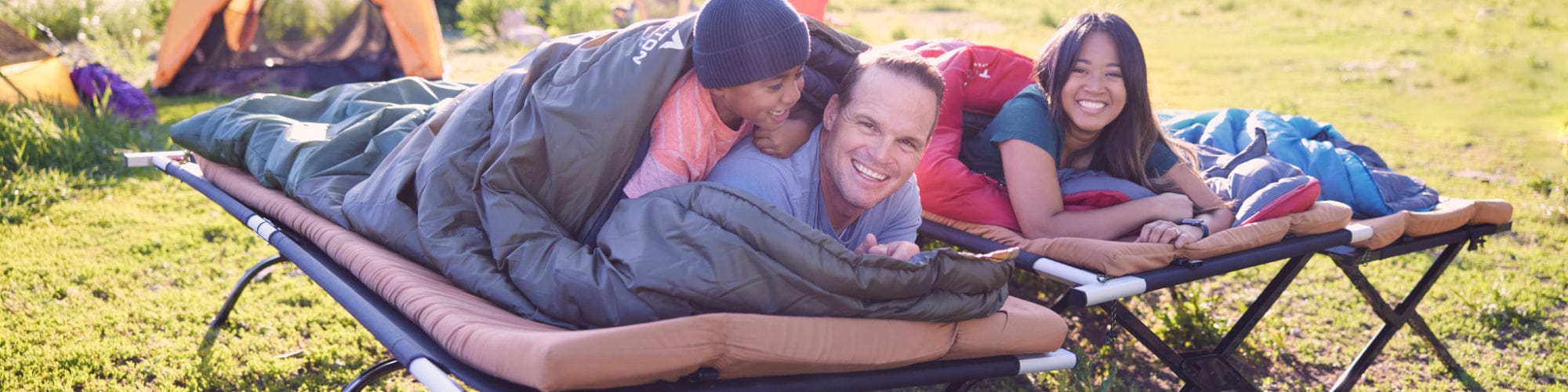 Two people lying on a camping cot and pads in a grassy field.