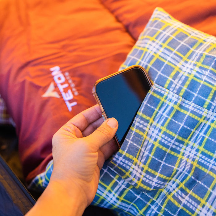 Hand holding a smartphone on a blue plaid sleeping bag with an orange shell material in the background