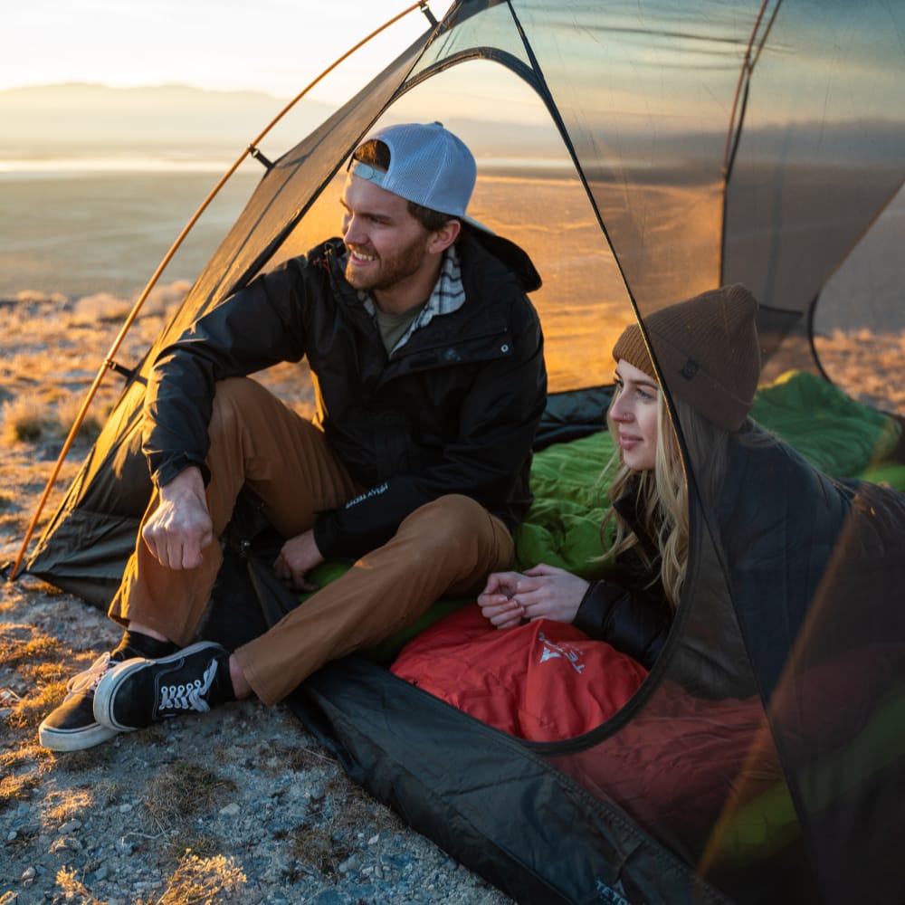Two people sitting inside the mesh of Mountain Ultra 2-person tent in a beach setting without the cover.