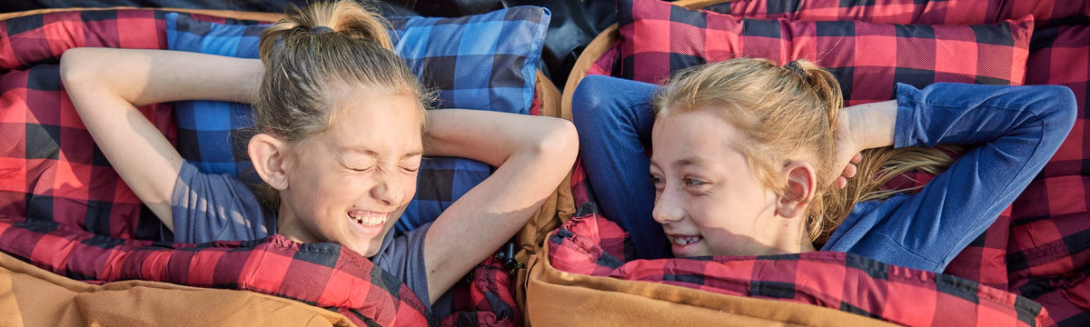 Two girls lying on buffalo plaid camp pillows, laughing together outdoors.