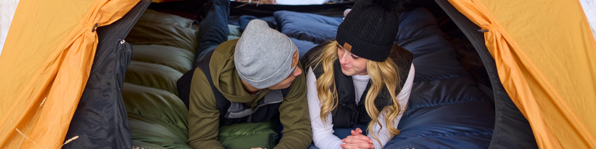 Two people inside a yellow tent, one wearing a beanie and the other a black hat lying on Green and Blue Response Sleeping Bags.