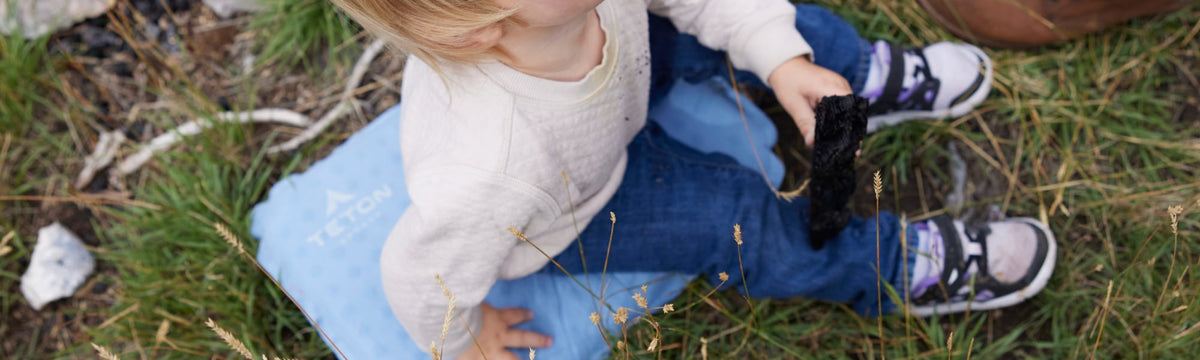 Toddler sitting on a blue TETON seat cushion in the grass by a campfire.