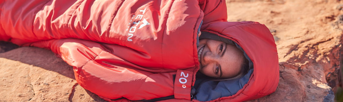 Close-up of a woman inside a red TETON Sports 20-degree mummy sleeping bag on red rock terrain.