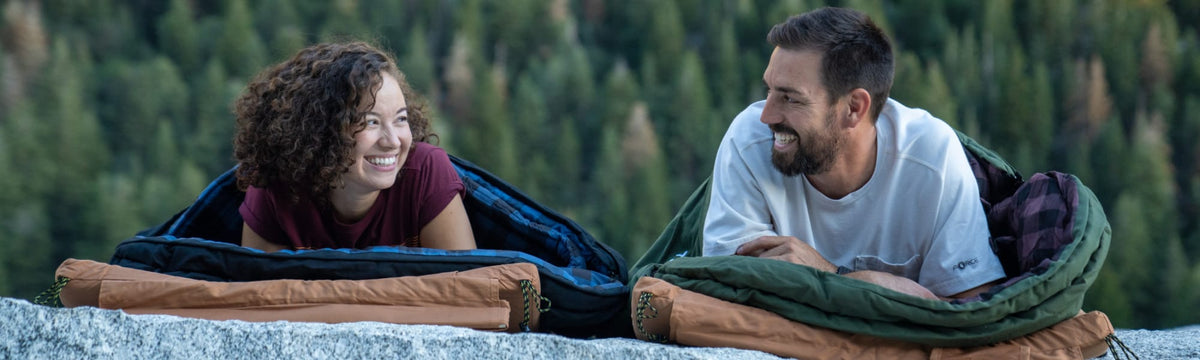 Man and woman laughing while lying in open mummy sleeping bags on a granite outcrop.