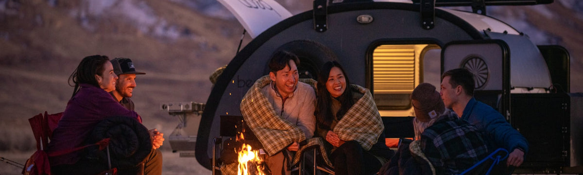 Friends wrapped in sleeping bags around a campfire beside a teardrop camper at dusk.
