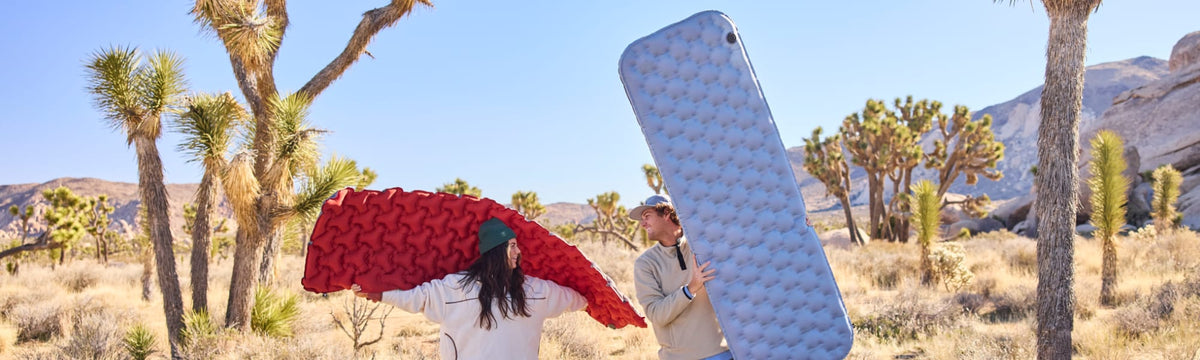 Couple playfully holding up a red inflatable sleeping pad and a blue self-inflating sleeping pad among Joshua trees.