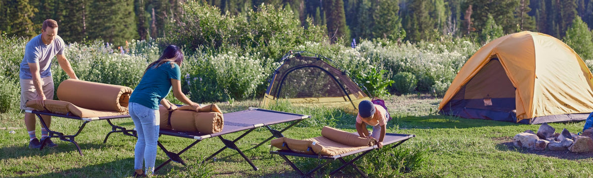 Family setting up sleeping pads on camping cots at a campsite with tents pitched nearby.