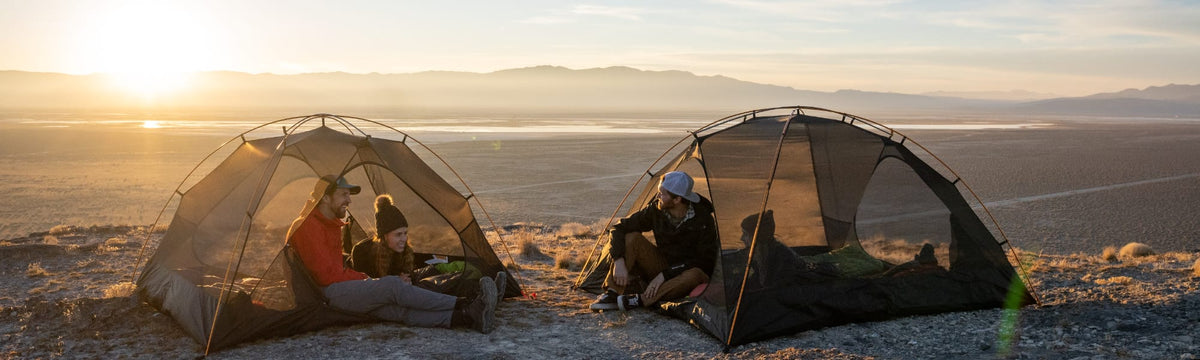 Two backpacking tents pitched on a desert overlook at sunrise, with hikers relaxing inside each one.