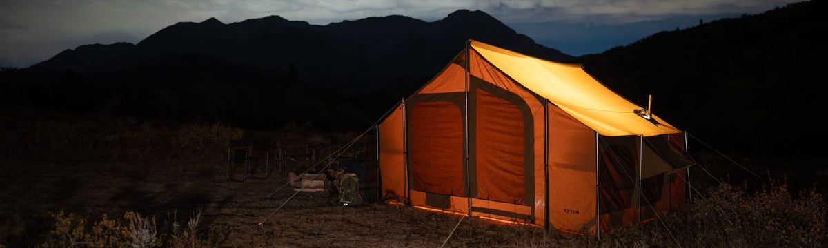 Canvas wall tent with a stove jack glowing orange at night against a mountain silhouette.