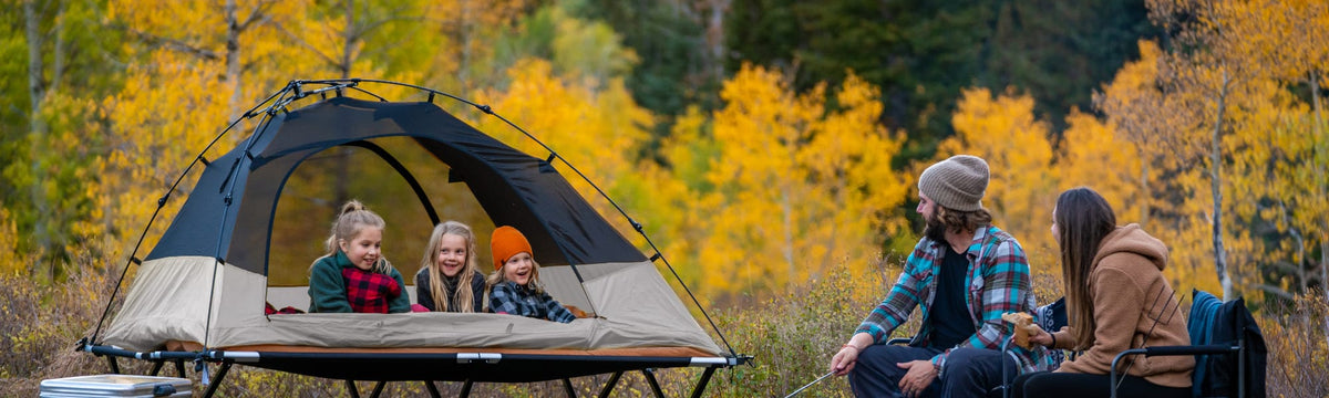 Three kids laughing inside a cot tent while parents sit by the fire in a golden aspen grove.