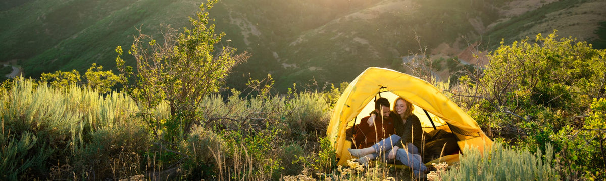 Couple sitting together inside a yellow dome tent overlooking a green mountain valley at sunset.